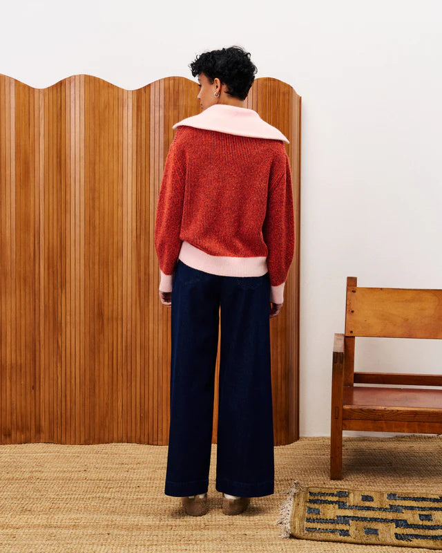 Person wearing a red sweater with a white collar and dark pants standing in front of a wooden screen.