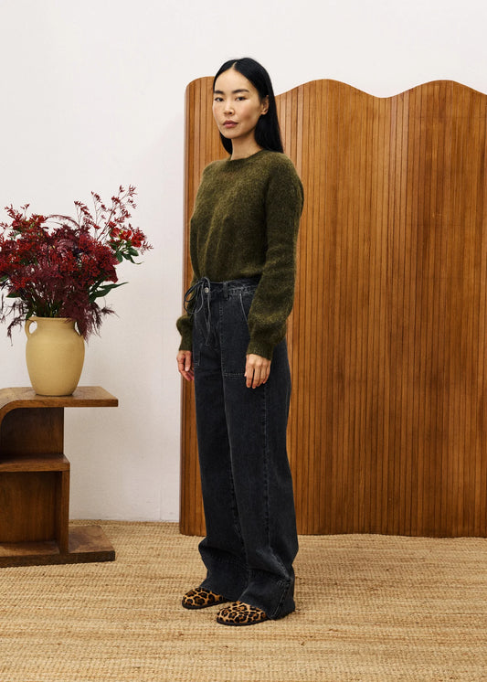 Woman in a green sweater and dark jeans standing in front of a wooden screen with a vase of flowers on a table.