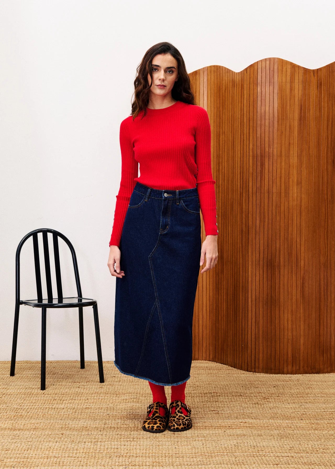 Woman wearing a red sweater and dark blue skirt standing in a room with a black chair and wooden wall.