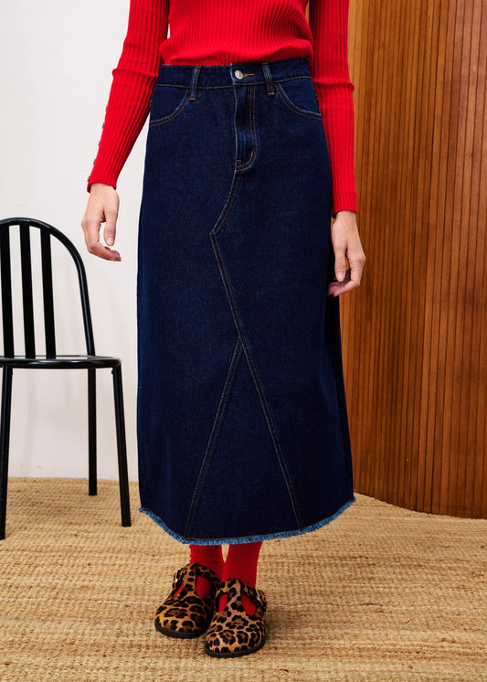 Person wearing a red sweater, blue denim skirt, and leopard print shoes in a room with a chair and wooden wall.