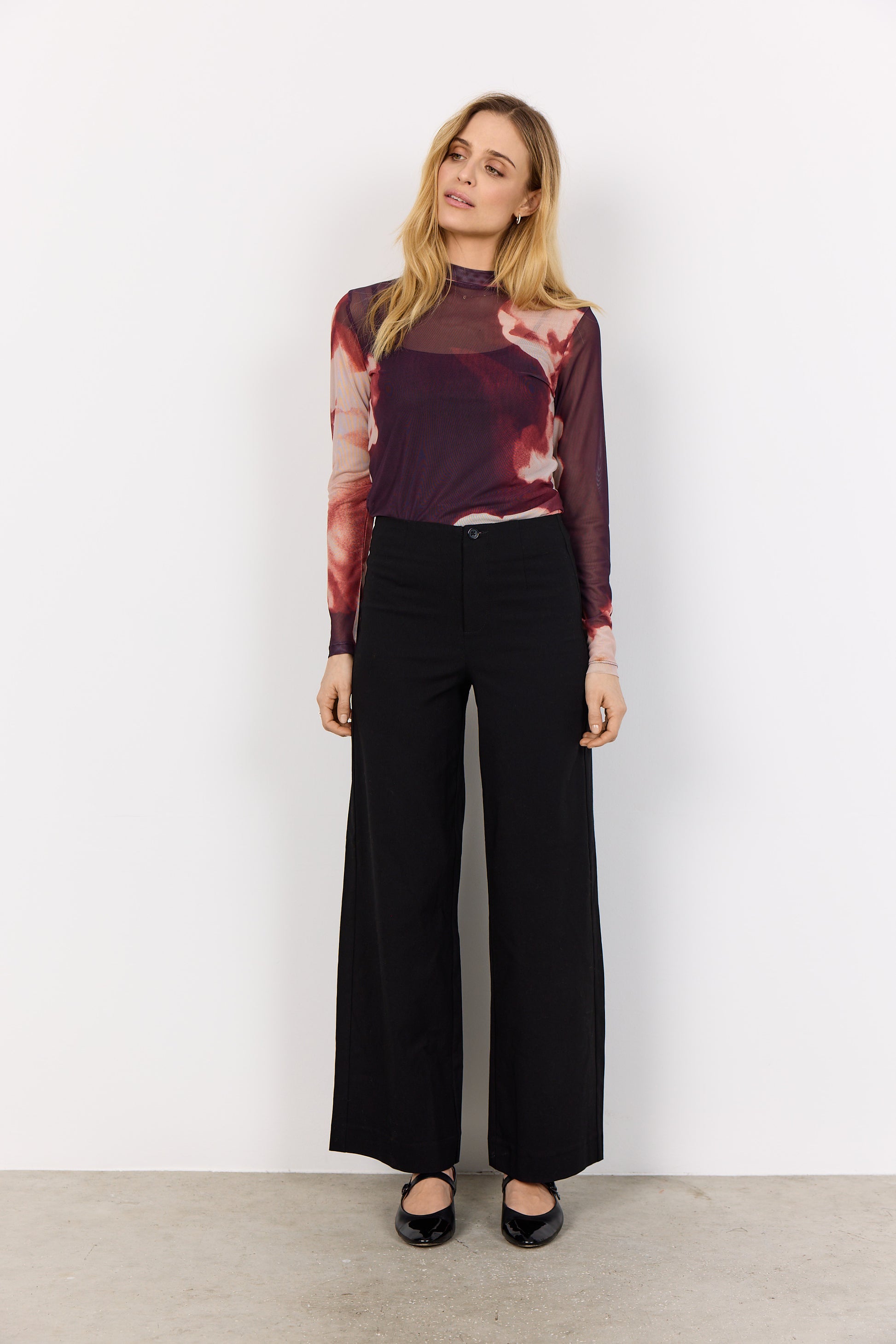 Woman wearing a patterned top and black pants against a white background