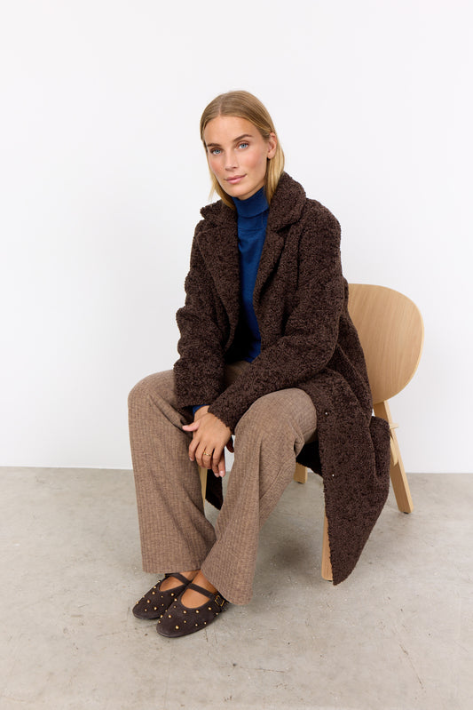 Woman sitting on a chair wearing a brown textured coat against a white background