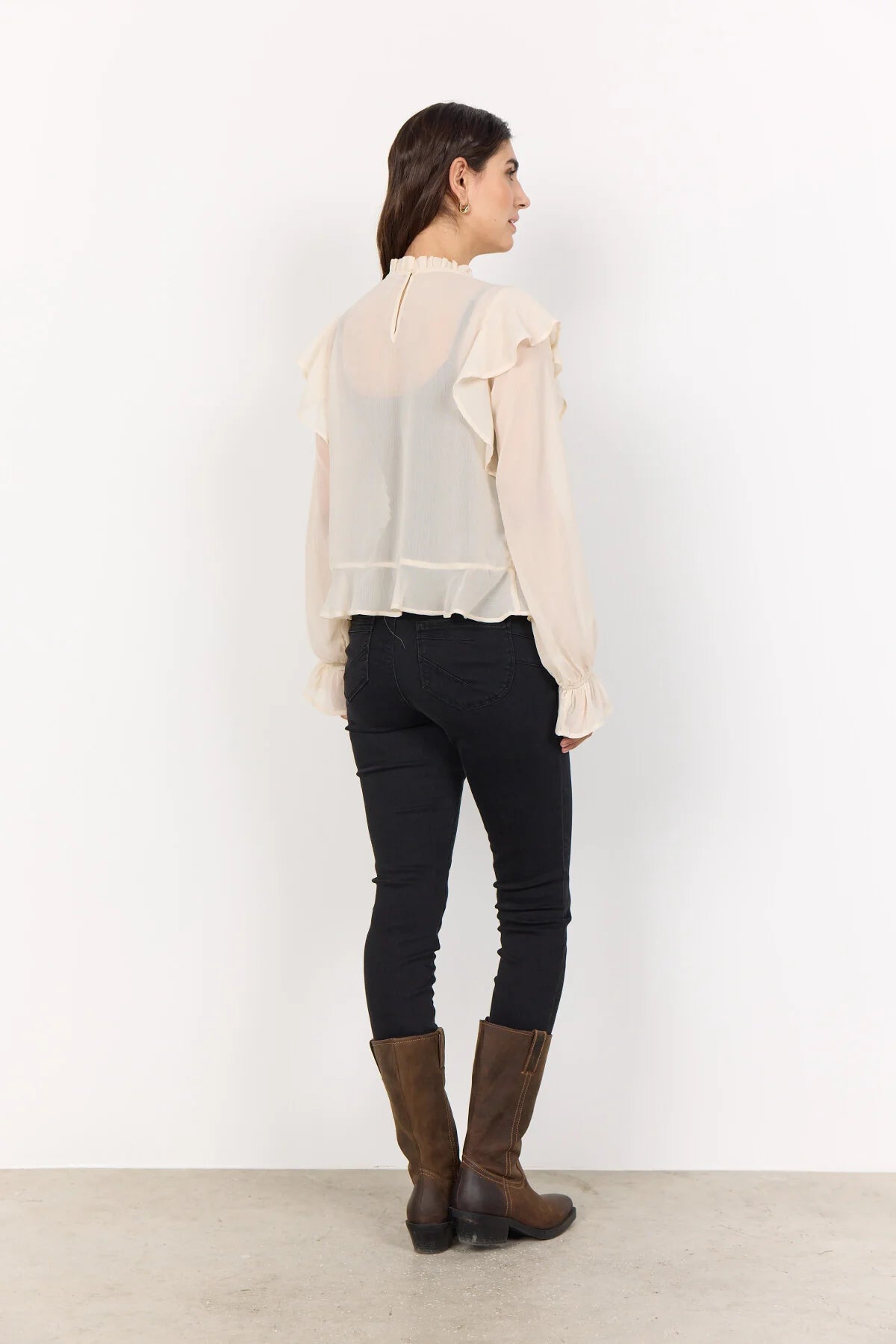 Woman wearing a cream blouse and black trousers and brown boots with a white background