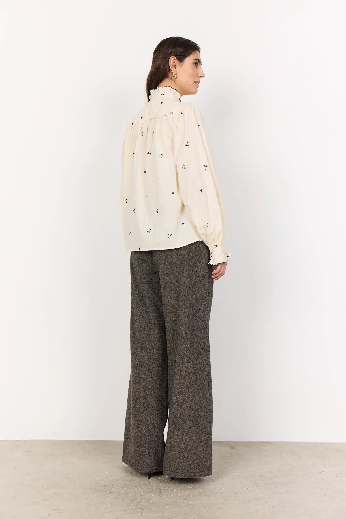 Woman wearing a cream blouse with black patterns and gray pants against a white background