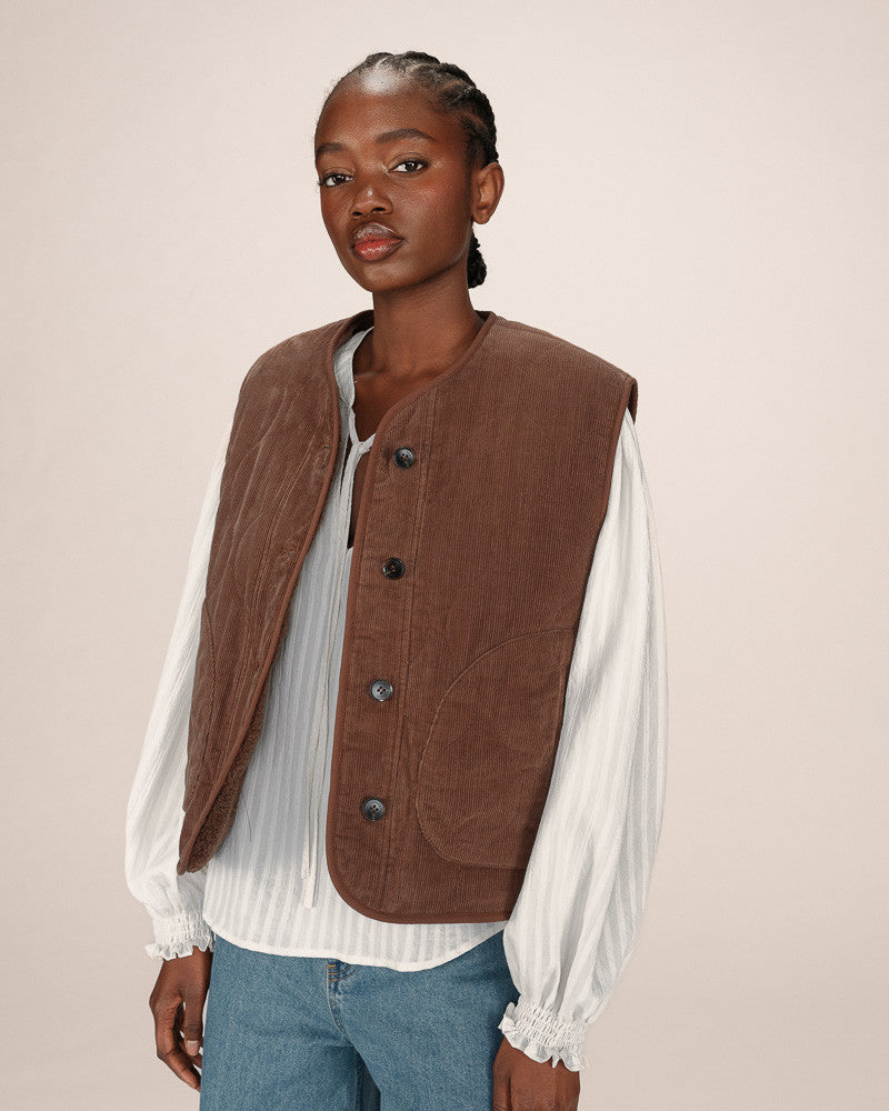 Woman wearing a brown vest over a white shirt and blue jeans on a beige background