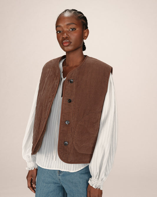 Woman wearing a brown vest over a white shirt and blue jeans on a beige background