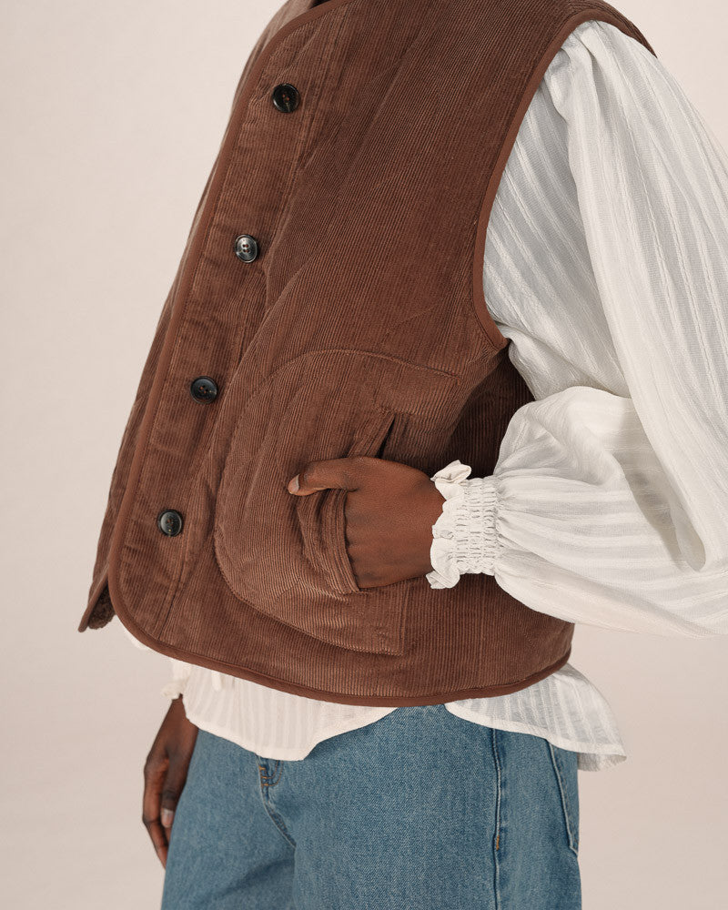 Brown corduroy vest over a white shirt and blue jeans on a beige background
