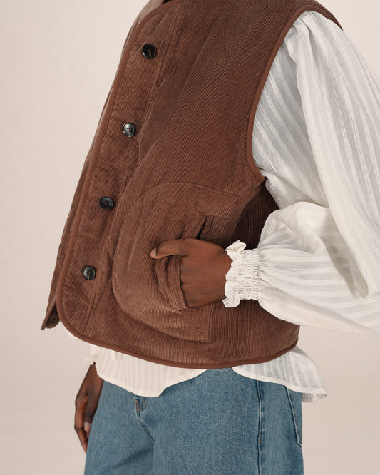 Brown corduroy vest over a white shirt and blue jeans on a beige background