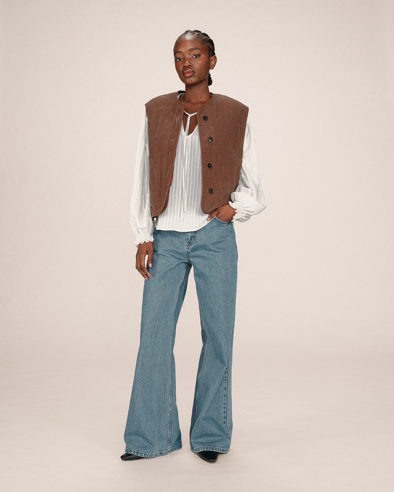 Person wearing a brown vest over a white blouse and blue jeans on a beige background
