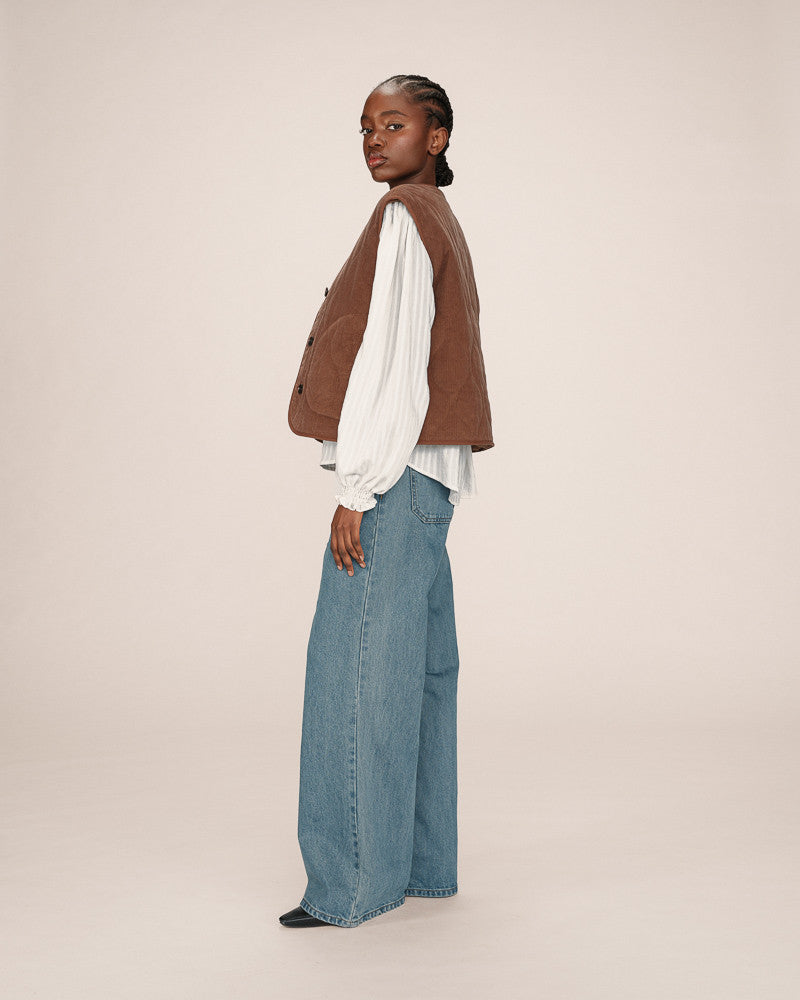 Woman wearing a brown jacket, white blouse, and blue jeans on a beige background