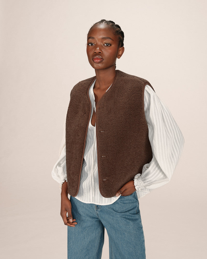 Woman wearing a brown vest over a white shirt and blue jeans on a beige background