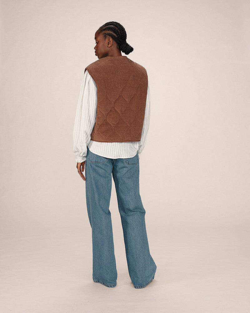 Person wearing a brown quilted vest over a white shirt and blue jeans on a beige background