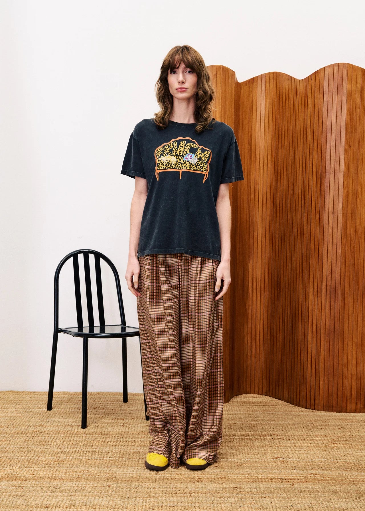 Person wearing a black t-shirt with a graphic design and plaid pants standing in a room with a wooden wall and black chair.