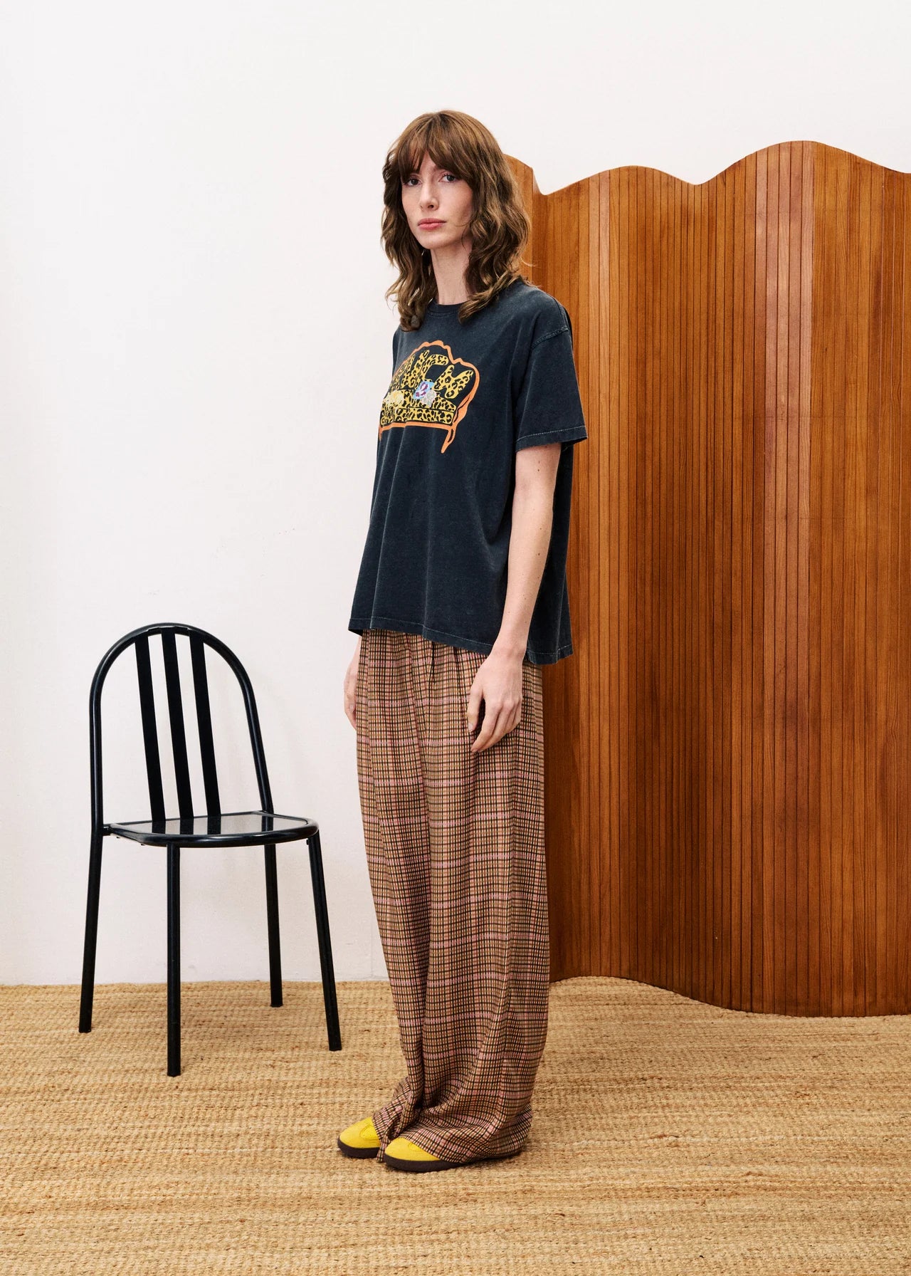 Woman wearing a black t-shirt with a graphic design and plaid pants standing in a room with a black chair and wooden wall.