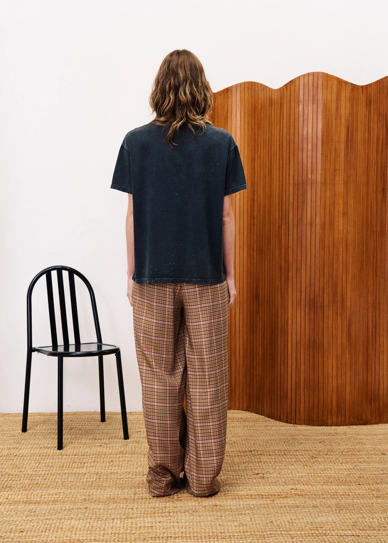 Person wearing a black t-shirt and plaid pants standing in front of a wooden screen with a black chair on a beige floor.
