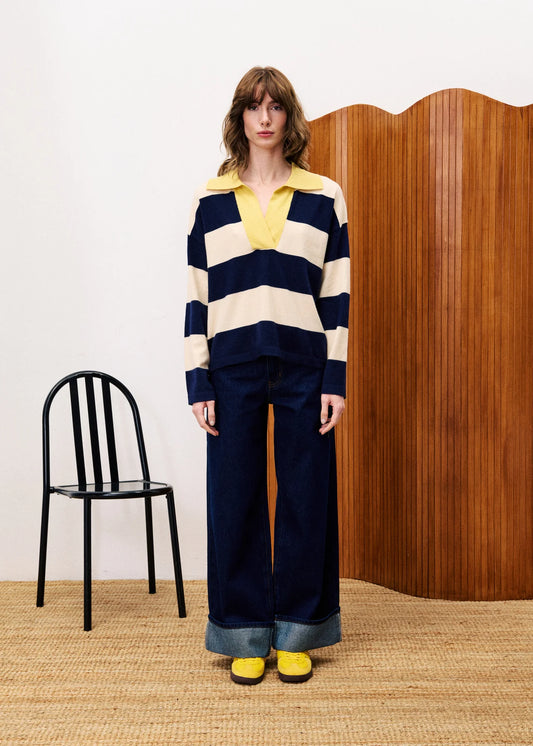 Model wearing a striped sweater and wide-leg pants with a yellow top, standing in a room with a black chair and wooden wall.