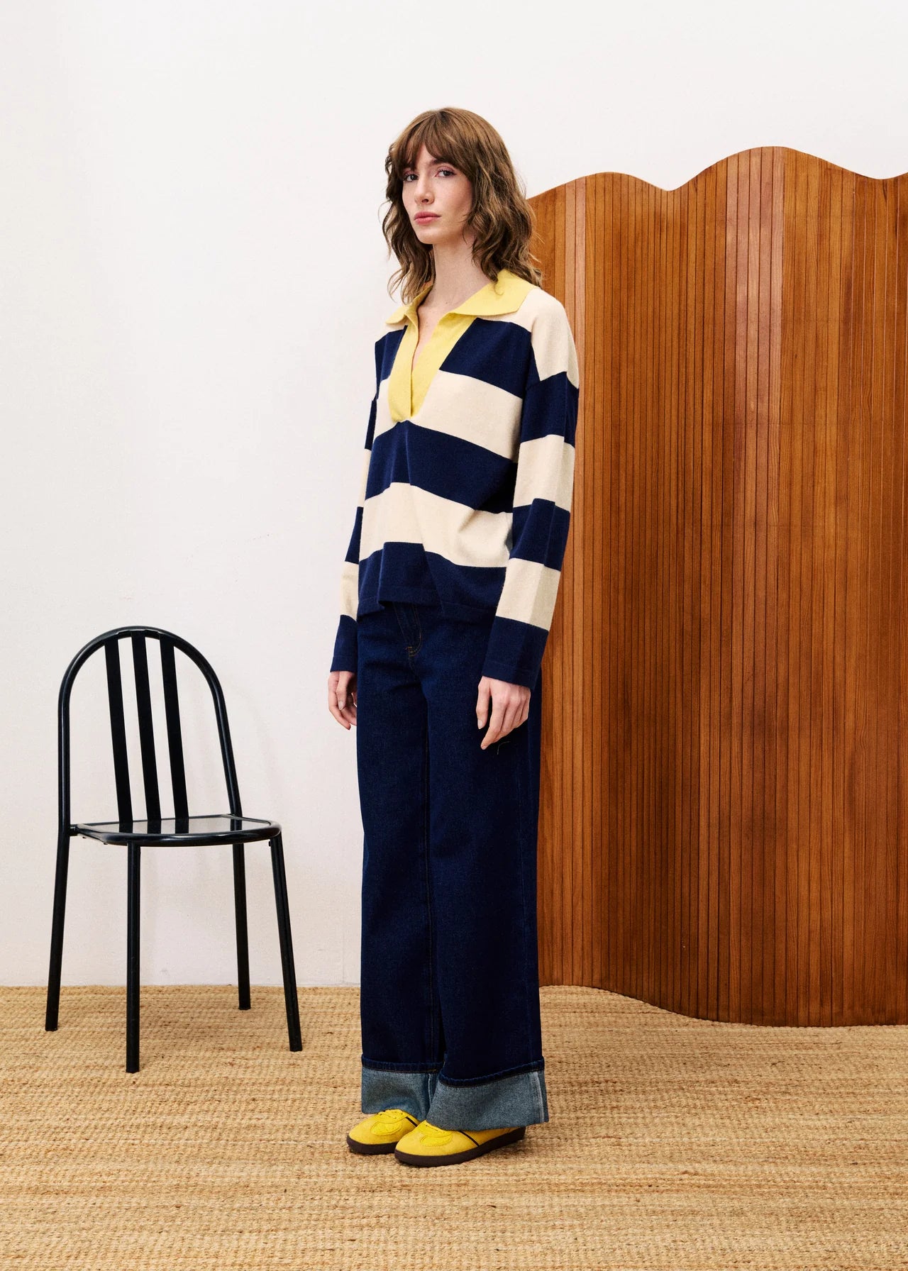 Woman wearing a striped sweater and wide-leg pants standing in a room with a black chair and wooden screen.