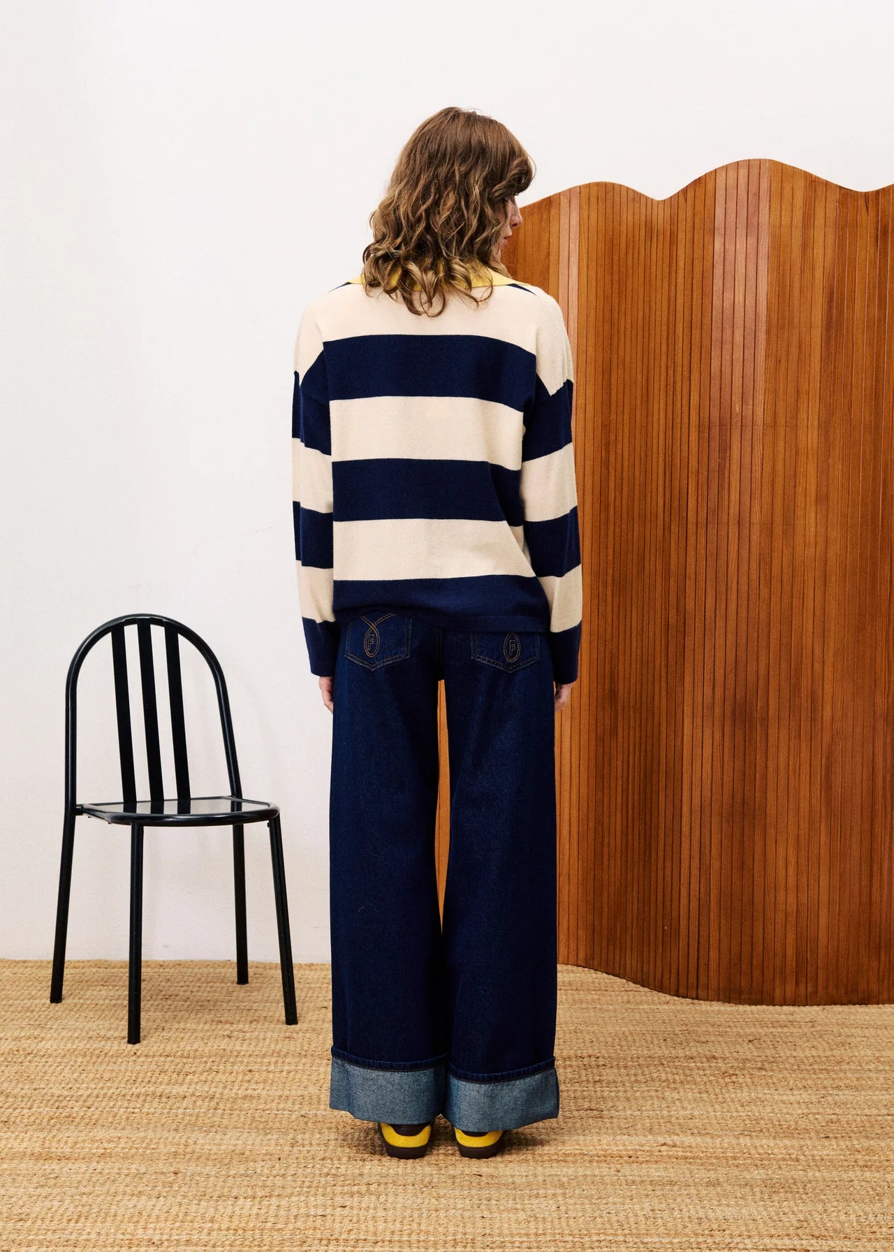 Person wearing a striped sweater and jeans standing in front of a wooden screen with a black chair to the left.
