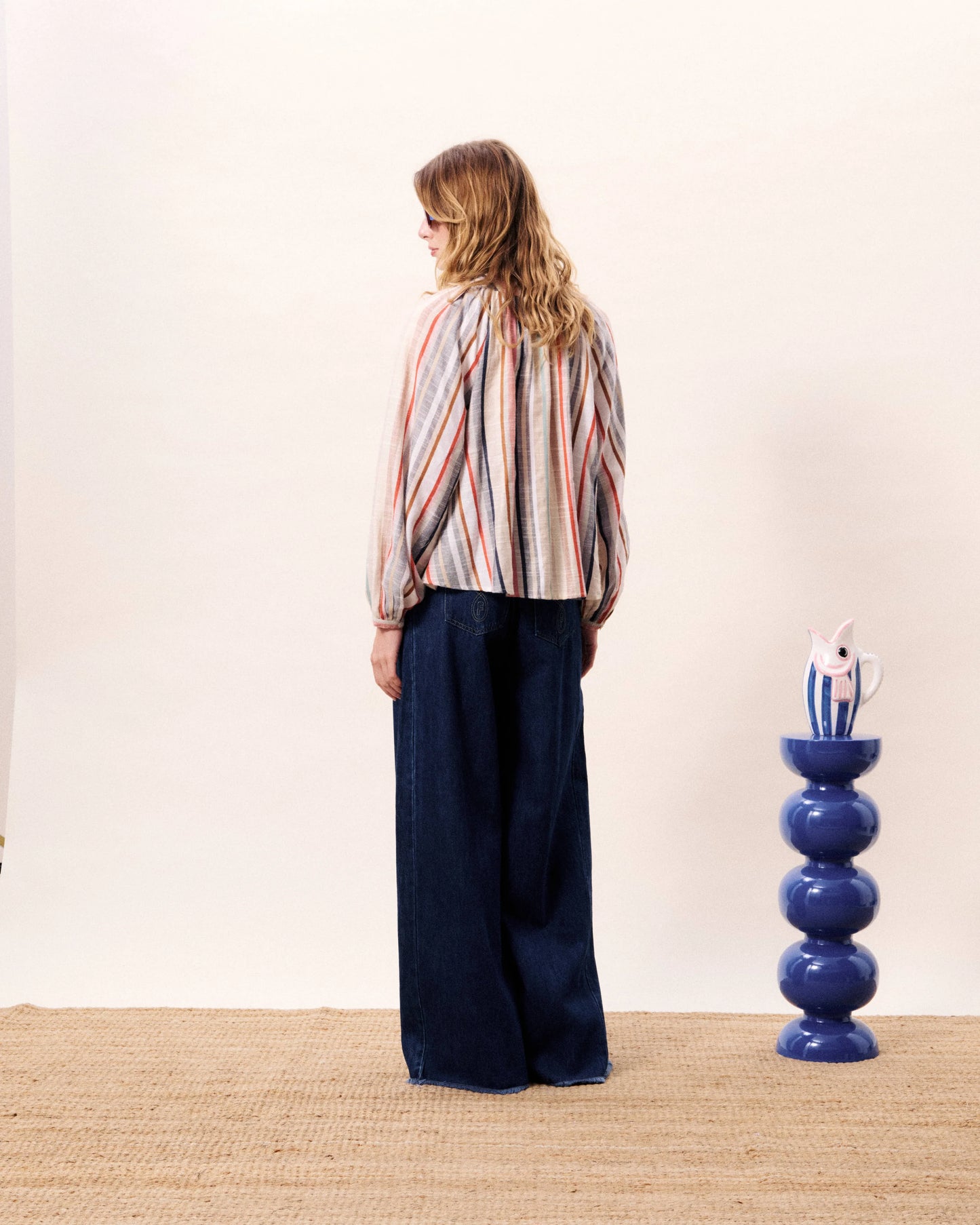 Person wearing a striped shirt and dark pants standing in front of a white wall with a blue decorative object.