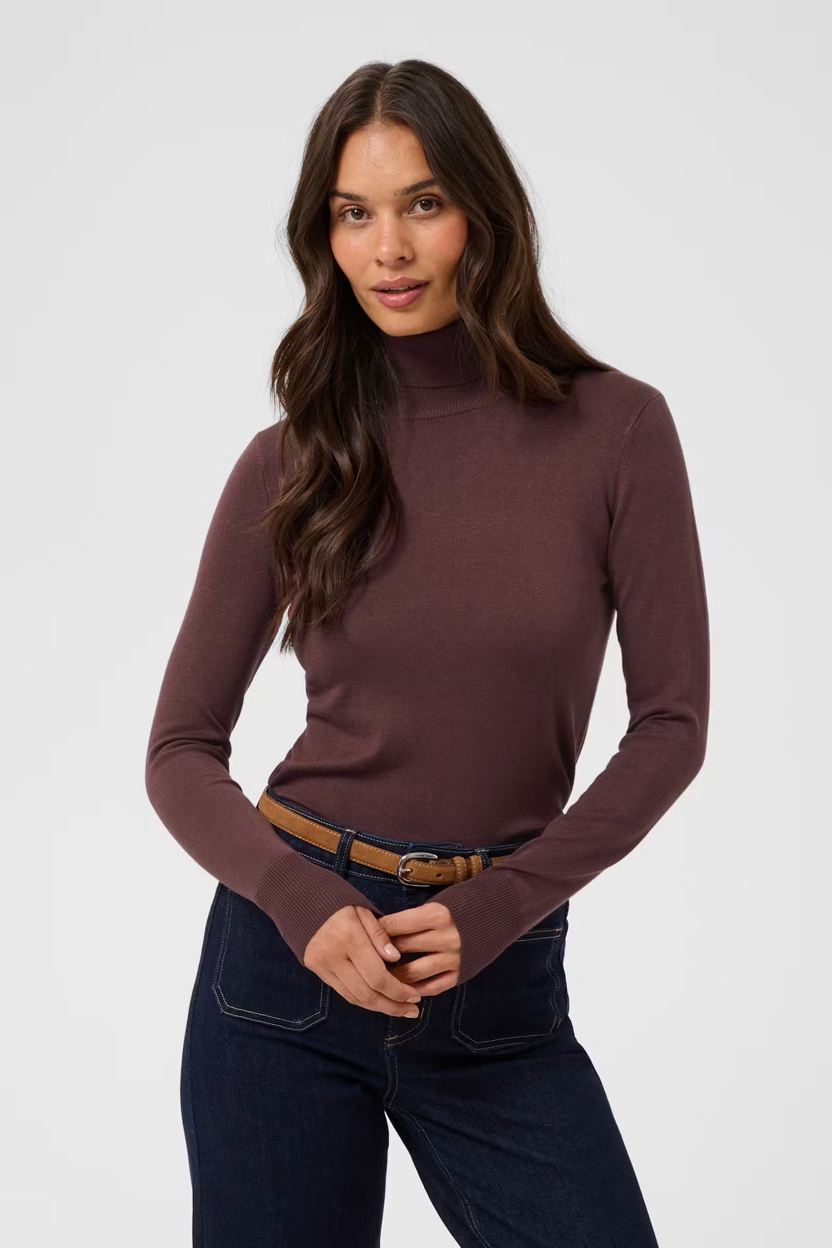 A woman modeling a fitted, long-sleeve mock neck turtleneck sweater in a muted plum-brown color, paired with dark blue jeans and a brown belt, against a light gray studio background.