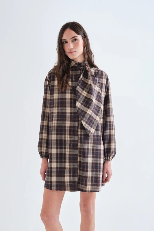 A woman modeling a long-sleeve plaid shirt dress with a high collar and a large draped tie detail at the neckline, shown against a plain light gray studio background.
