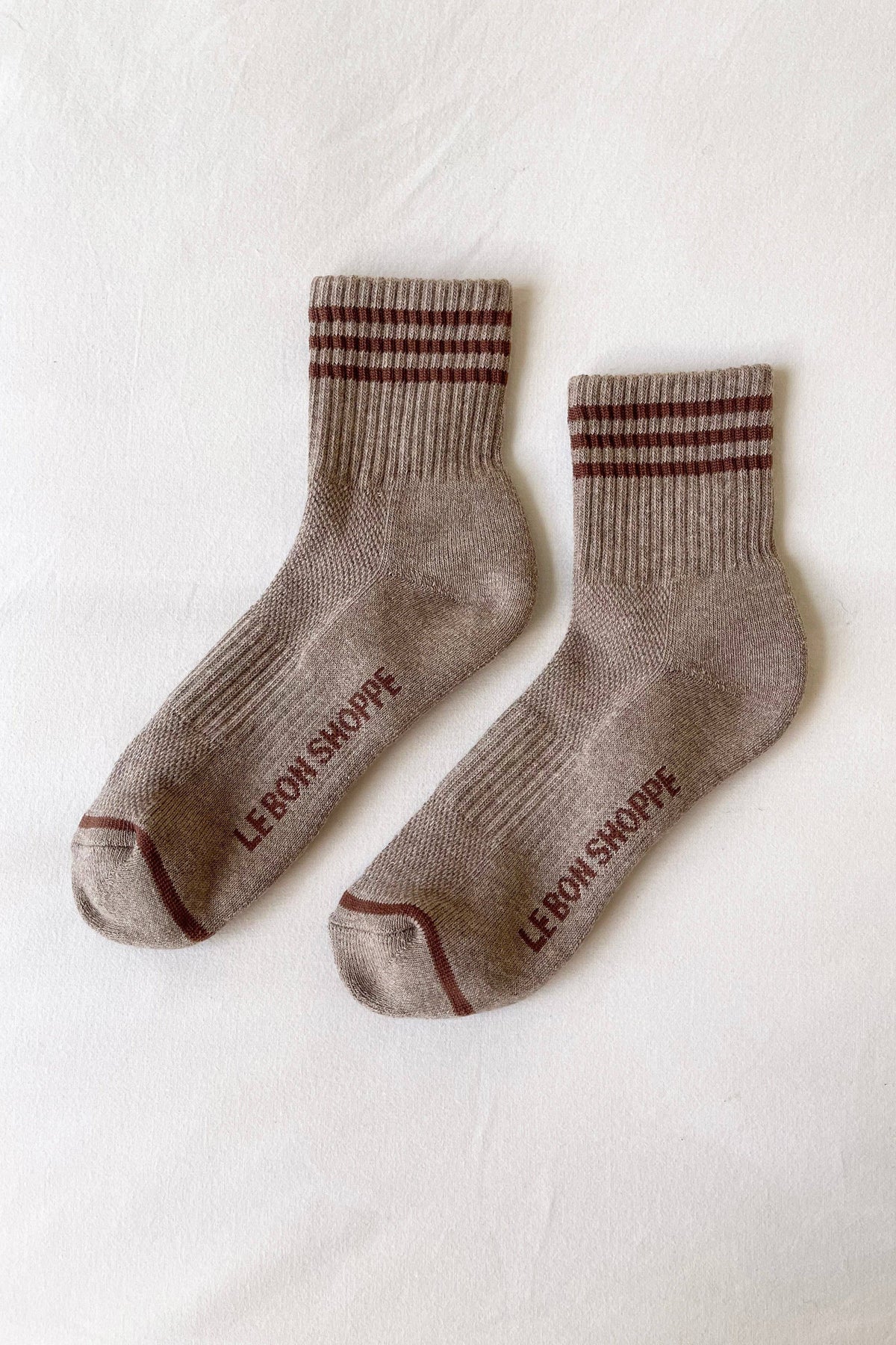 A pair of beige crew socks with dark burgundy stripes on the cuff and heel, featuring the brand name 'LERON SKOPPE' printed in burgundy on the sole, laid flat on a white textured fabric background.