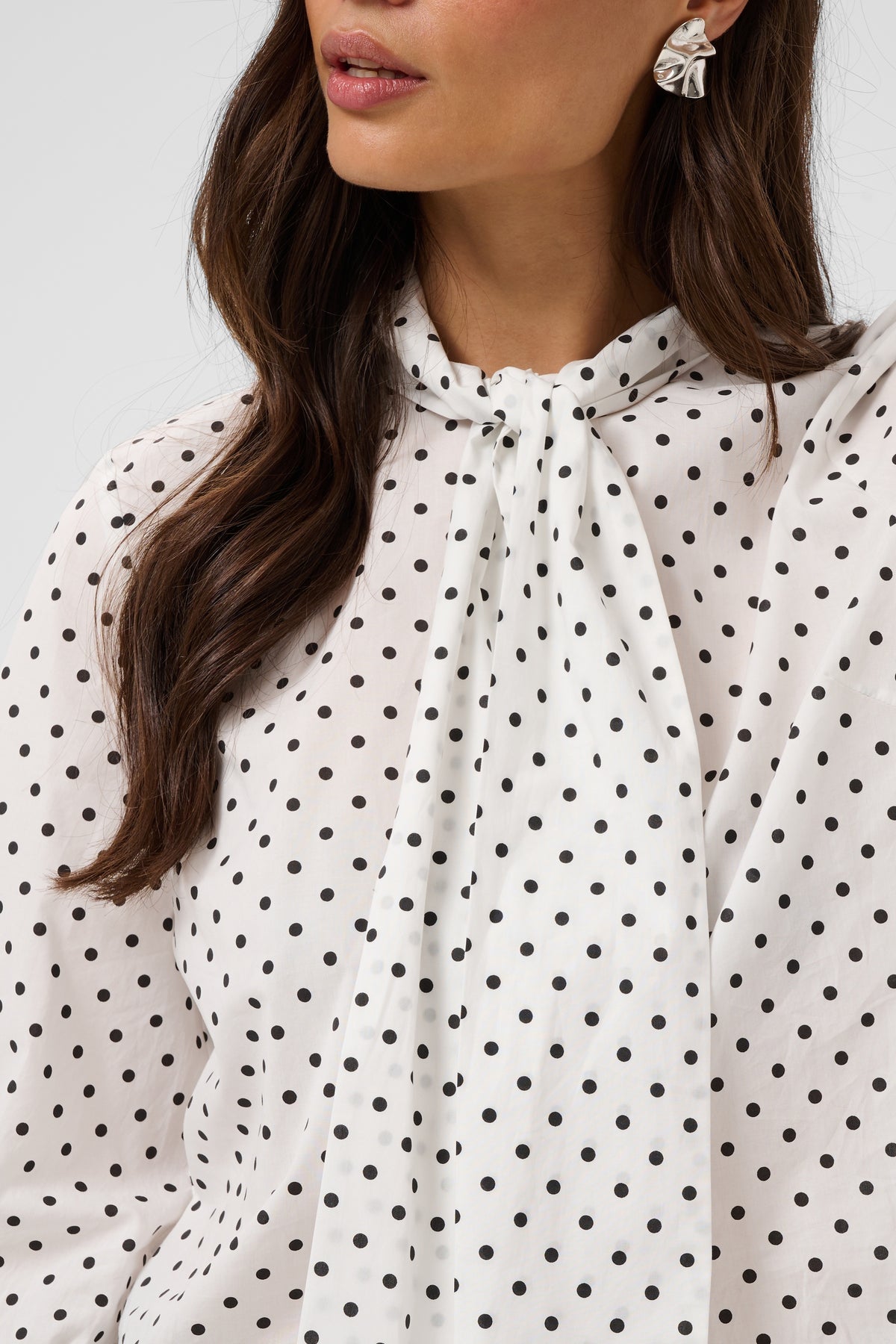 A close-up of a woman wearing a white long-sleeve blouse with a black polka dot pattern and a high neck tie detail.