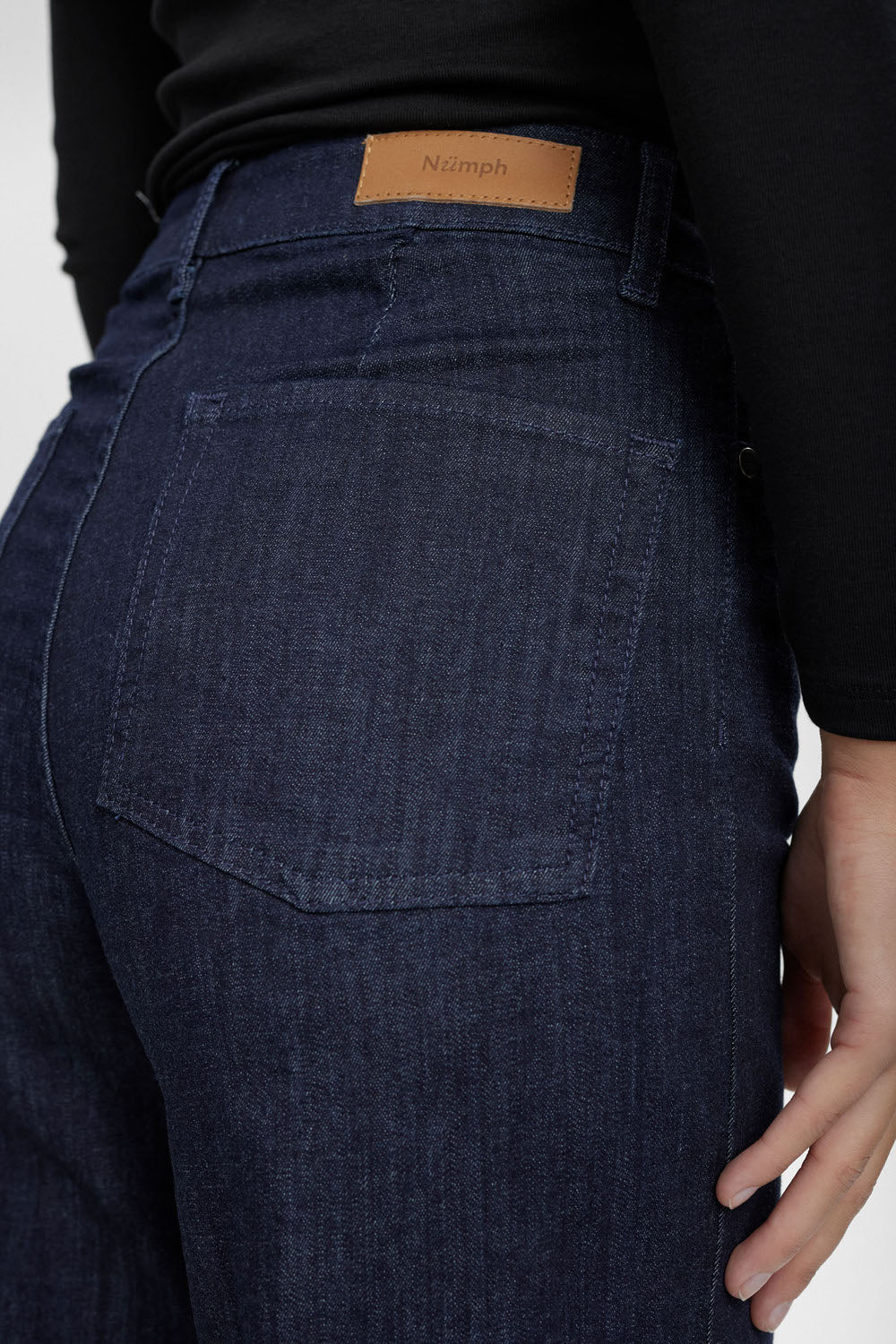 Close-up of the back waistband and pocket of dark blue denim jeans worn by a model, showing a tan leather patch with the brand name 'Nimph'.