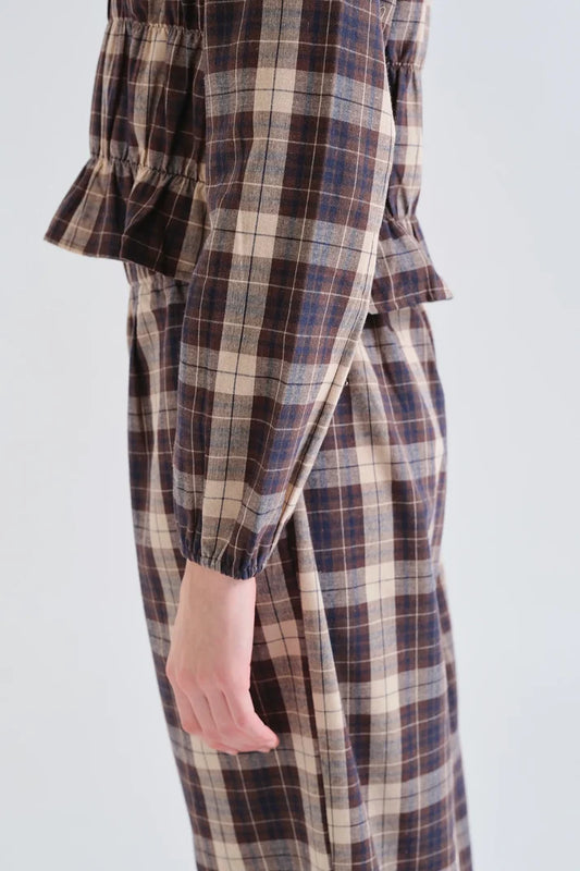 A close-up side view of a person wearing a plaid long-sleeve dress or jumpsuit in brown, beige, and navy tones, with a ruffled sleeve and gathered waist detail, against a plain light gray background.