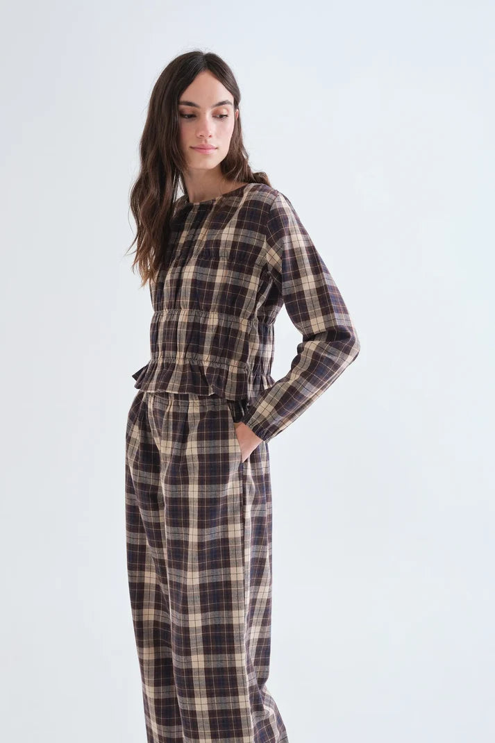 A woman modeling a matching plaid long-sleeve top and wide-leg pants set in a dark brown, beige, and black tartan pattern, set against a plain light gray studio background.