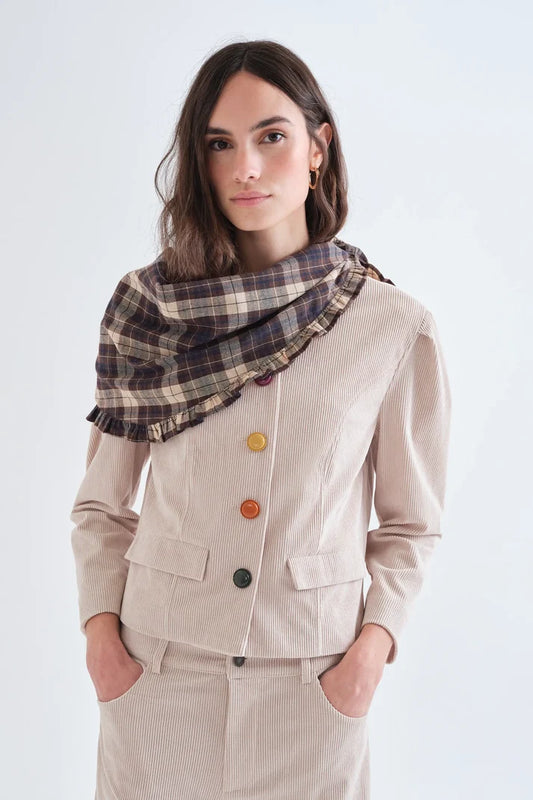 A woman wearing a beige double-breasted jacket with colorful buttons and a dark brown plaid scarf, photographed against a light gray studio background.