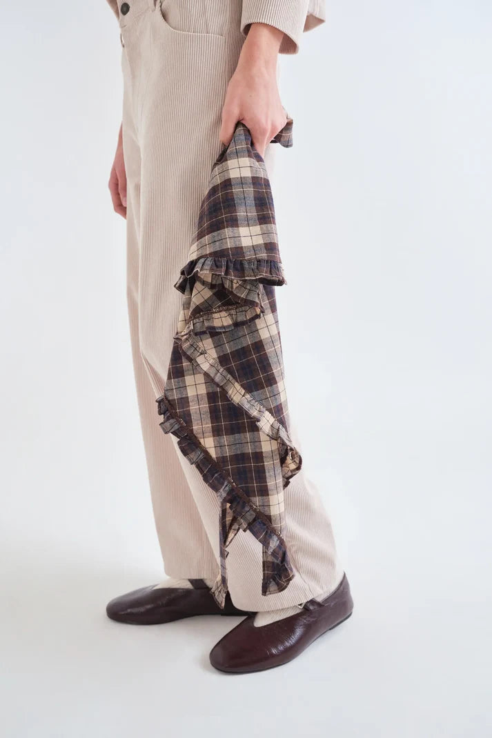 A person wearing beige wide-leg trousers and dark brown leather shoes holds a folded plaid shirt in a neutral studio setting.