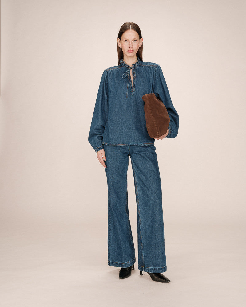 Woman wearing a blue denim outfit holding a brown clutch against a beige background