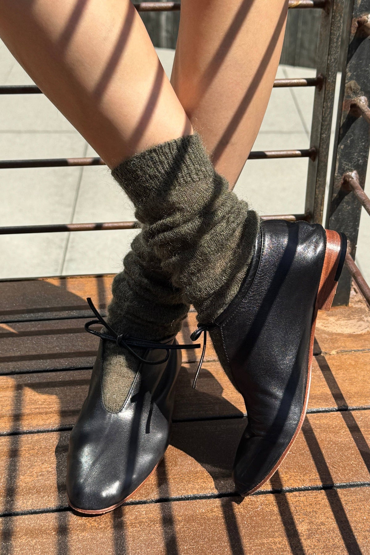 A person wearing black leather lace-up ankle boots with dark olive green knit cuffed socks, sitting on a wooden deck with metal railings in the background.