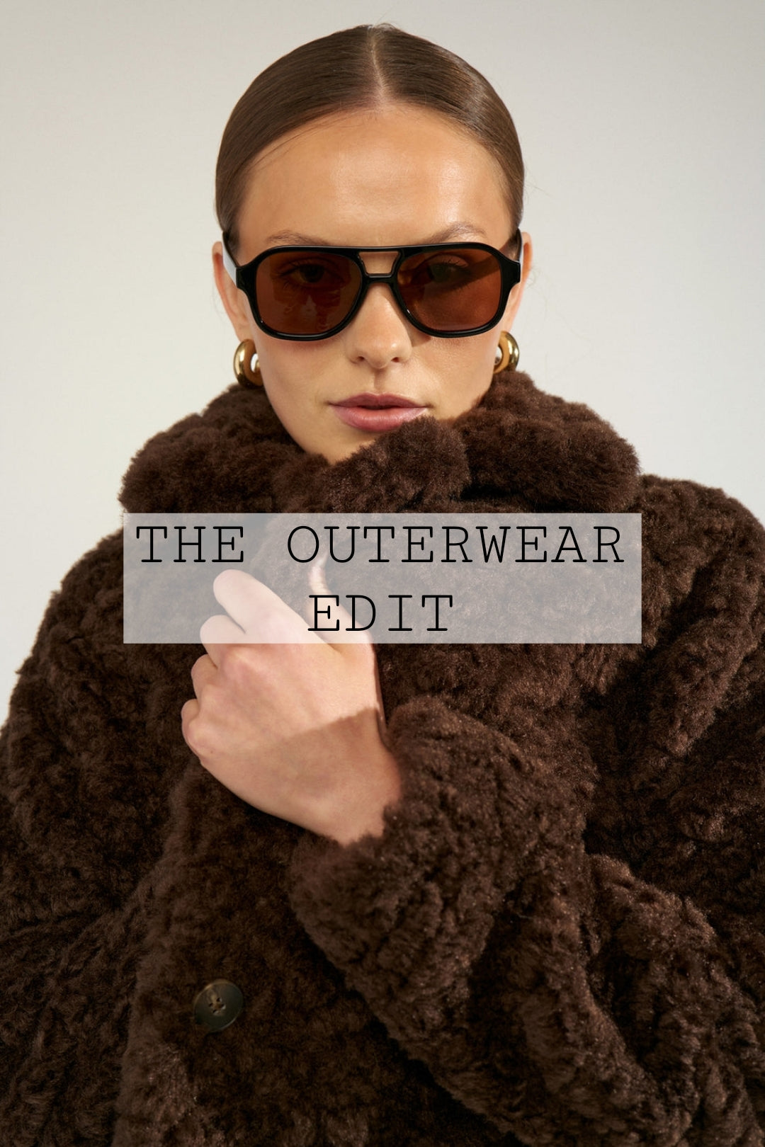 A model wearing a luxurious dark brown textured faux fur coat, black sunglasses, and gold hoop earrings, photographed against a neutral light gray background.