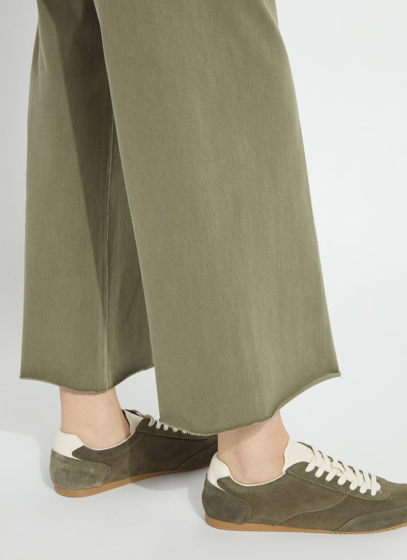 Close-up of a person wearing wide-leg olive green trousers and olive green suede sneakers with cream laces and white heel tabs, standing against a light gray studio background.