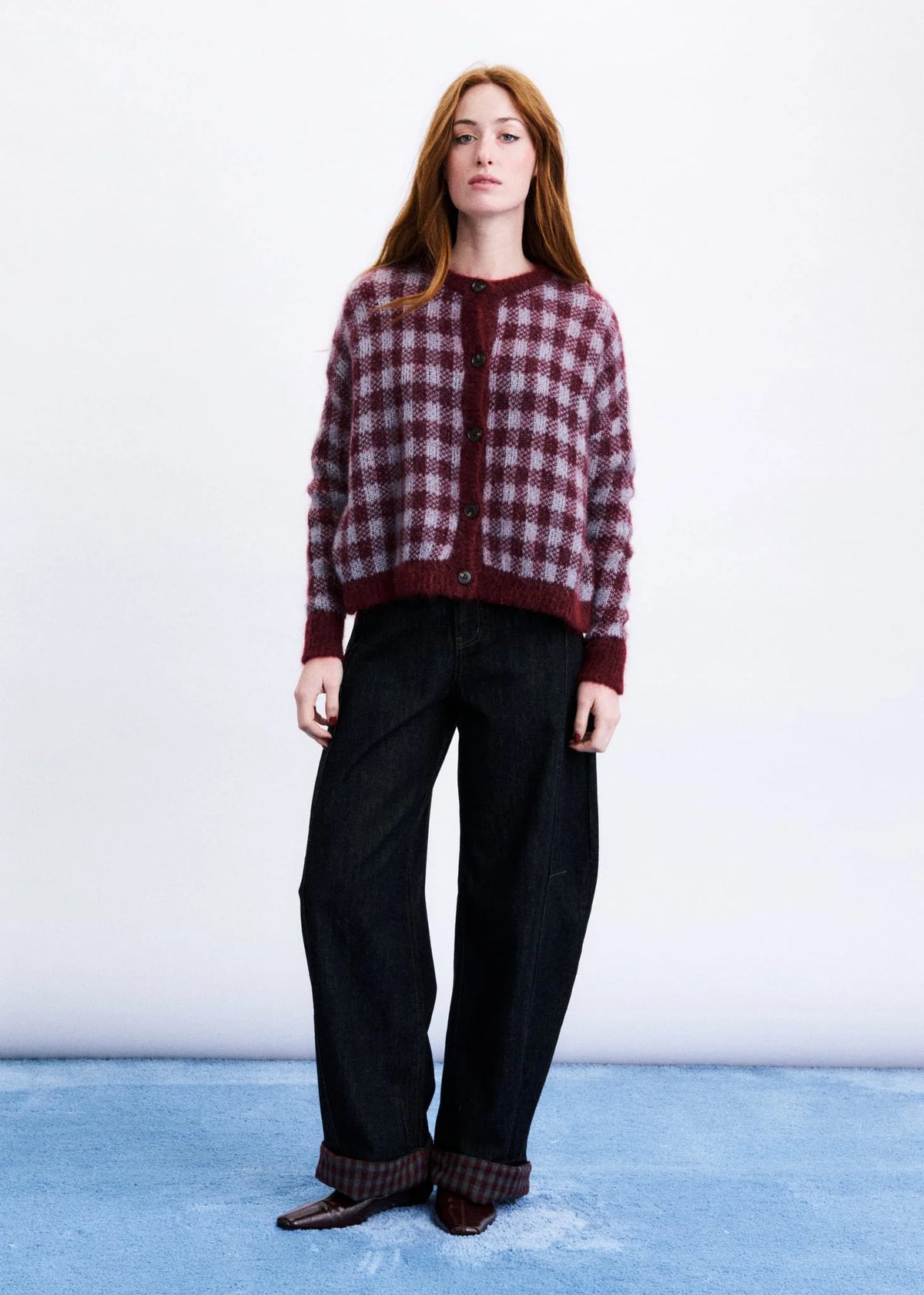 A woman stands against a white studio wall wearing a burgundy and dark red checkered cardigan, loose black wide-leg jeans, and brown shoes.