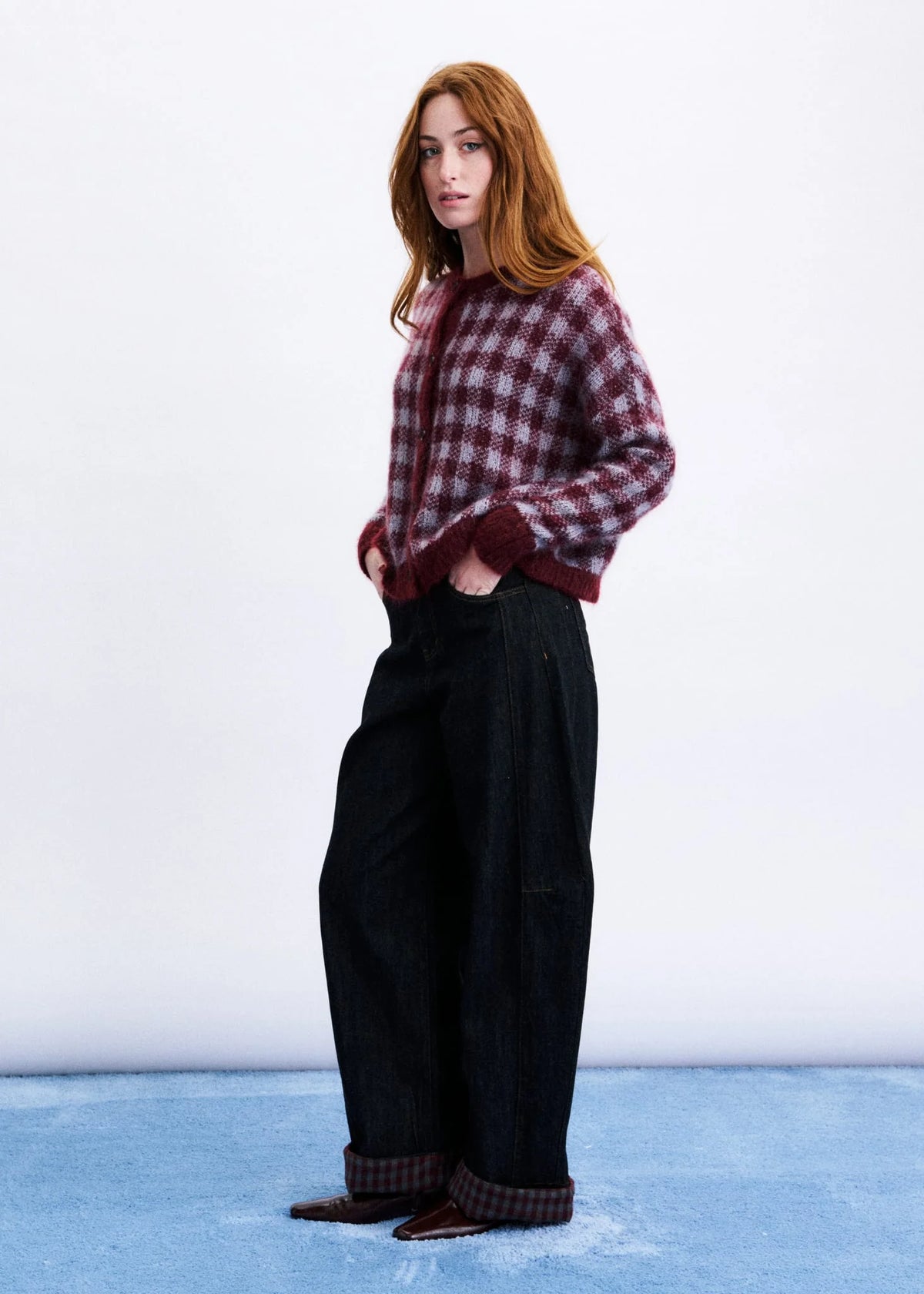 A woman stands in a studio wearing a burgundy and dark red plaid button-up shirt, wide-leg black trousers, and brown shoes with matching plaid cuffs.