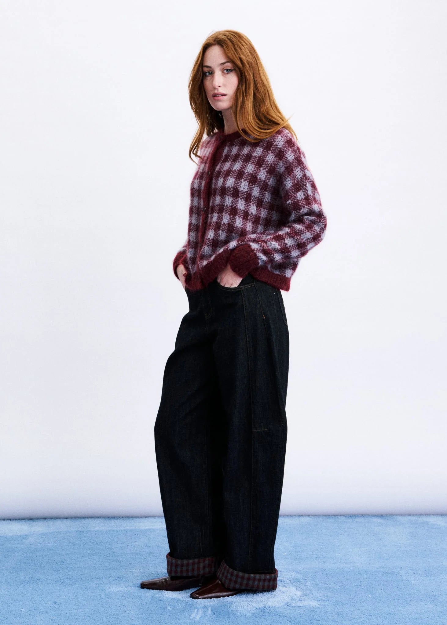 A woman stands in a studio wearing a burgundy and dark red plaid button-up shirt, wide-leg black trousers, and brown shoes with matching plaid cuffs.