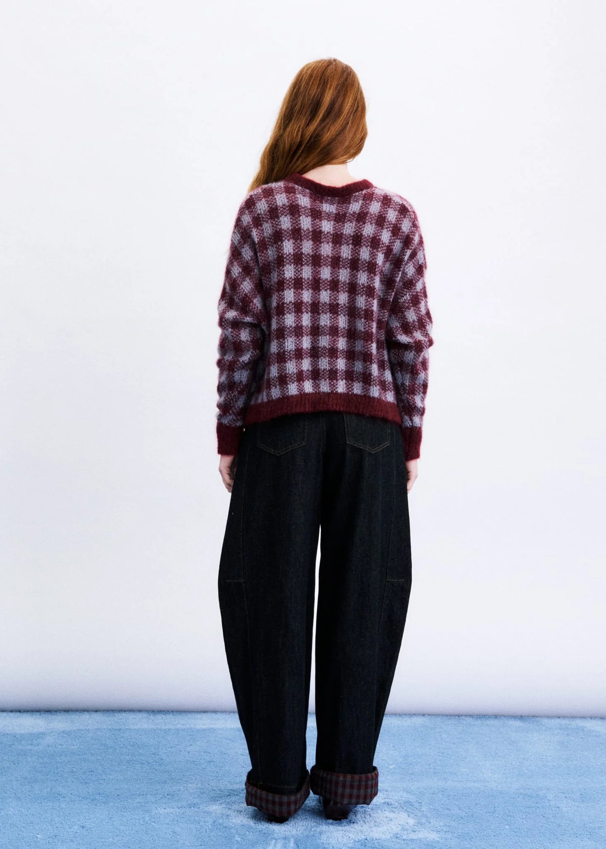 A woman viewed from behind wearing a burgundy and black plaid knit sweater, dark wide-leg jeans, and matching plaid socks, standing against a white wall and blue floor.