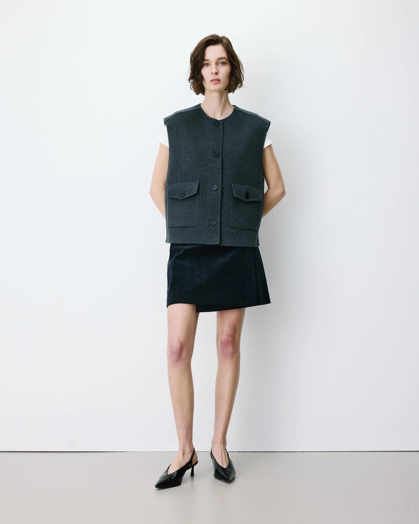 Woman wearing a dark vest over a white shirt and black skirt against a white background
