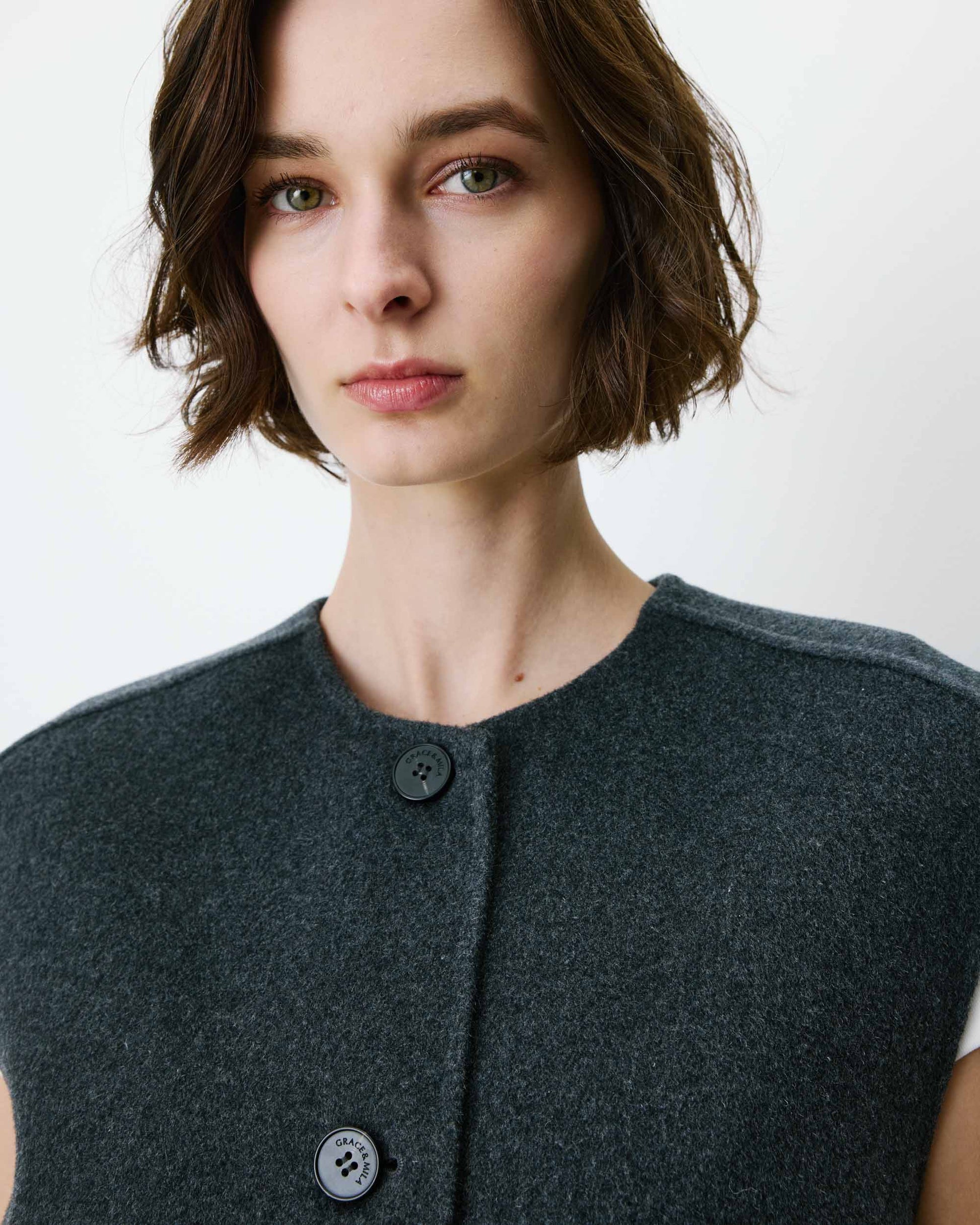 Woman wearing a dark gray sleeveless coat with buttons on a white background