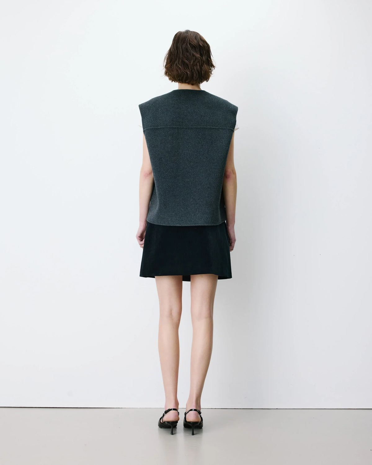 A model viewed from the back wearing a dark gray sleeveless knit vest over a black mini skirt, paired with black heeled sandals, set against a plain white studio wall.