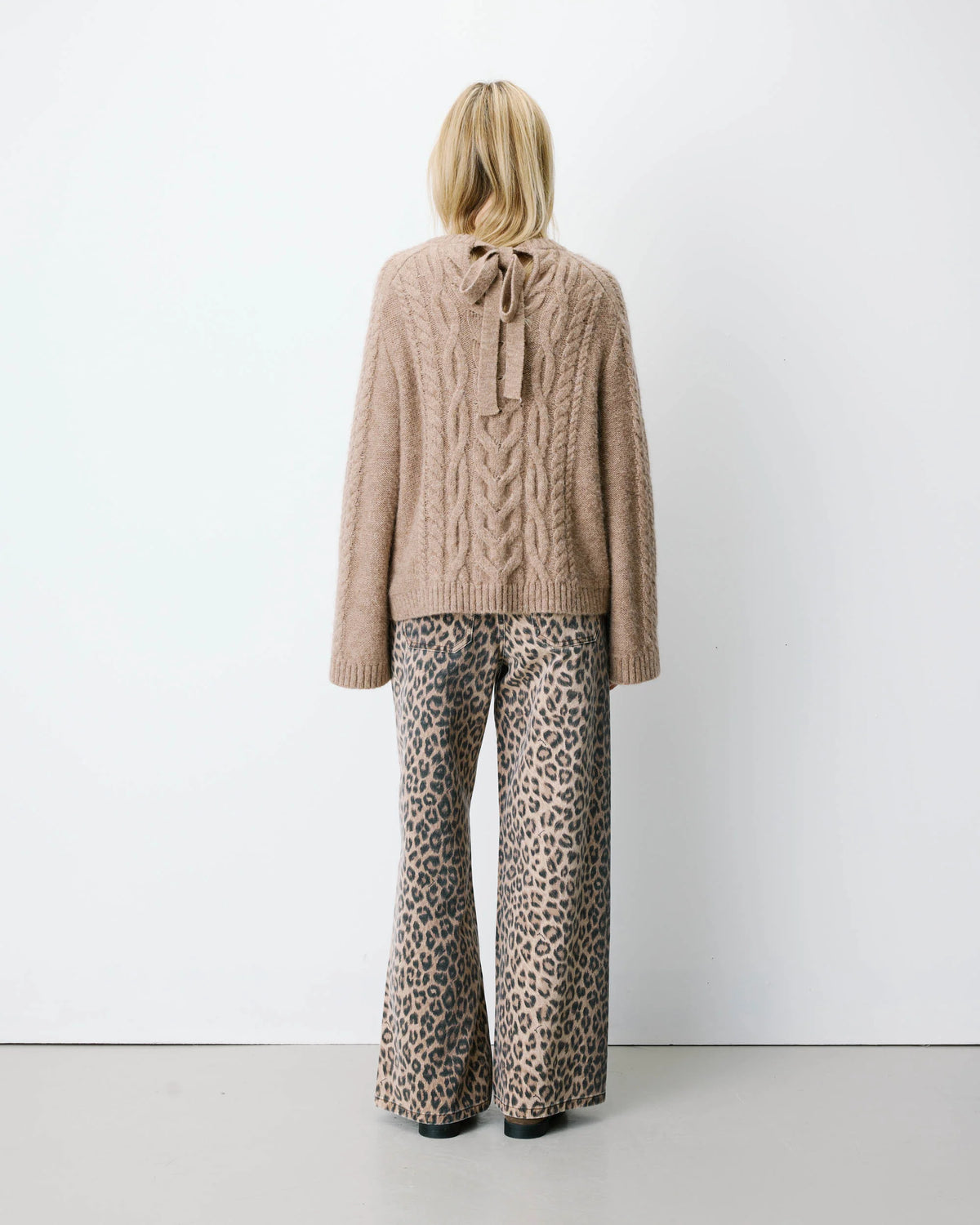 A model viewed from the back wearing a beige cable-knit sweater with a tie detail at the back neckline and wide-leg leopard-print pants, standing against a plain white studio wall.