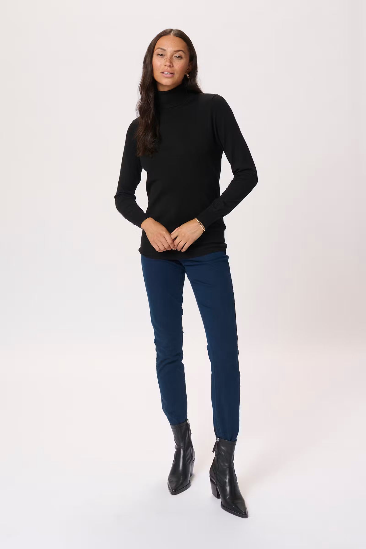 A woman modeling a black long-sleeve mock neck top, dark blue skinny jeans, and black ankle boots against a plain white studio background.