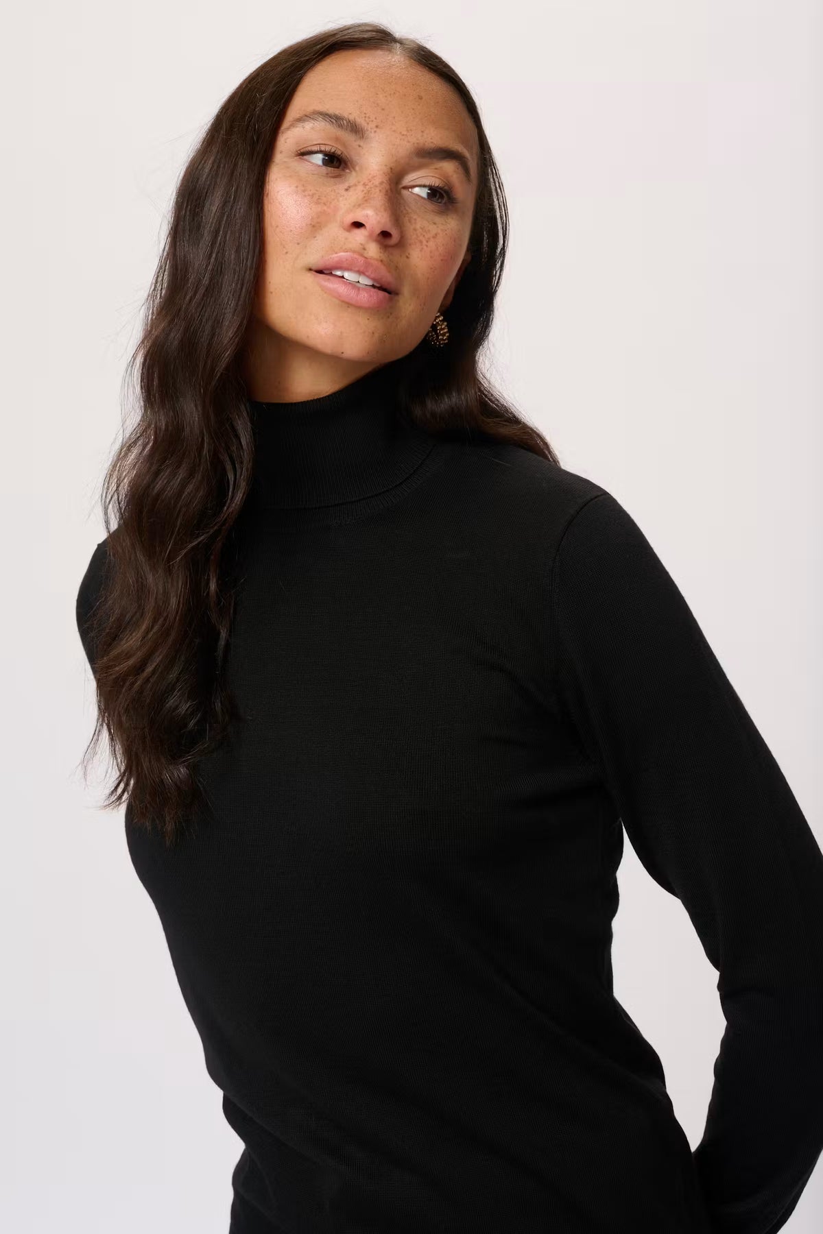 A woman with long dark brown hair wearing a black long-sleeve mock neck turtleneck top, photographed against a plain light gray studio background.