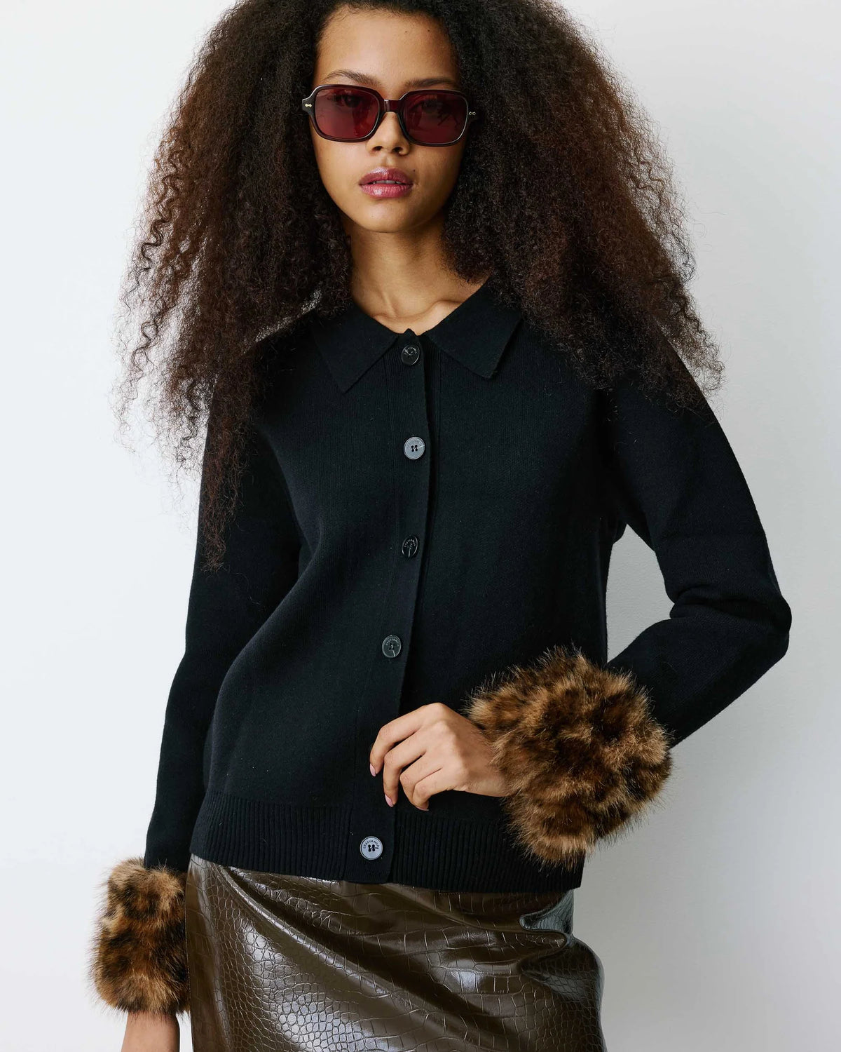 A model with curly hair wearing a black knit button-front cardigan with brown faux-fur trimmed cuffs, paired with a brown faux-leather skirt and dark sunglasses, against a light gray studio background.