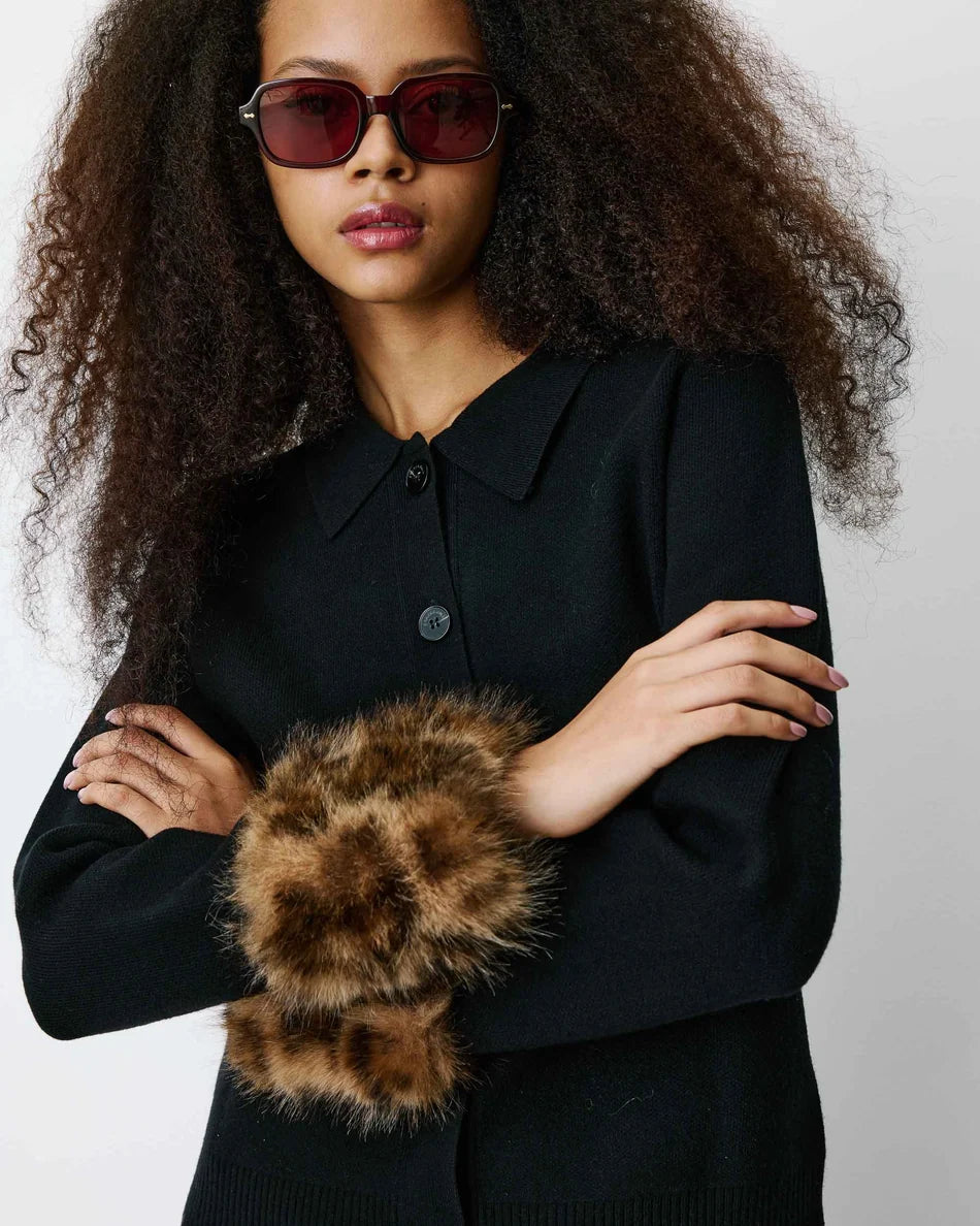 A model with voluminous curly hair wearing a black knit button-front top and a brown faux-fur wrist warmer, styled with dark sunglasses against a light gray background.