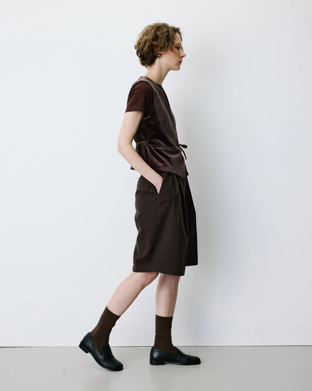A model is shown from the side wearing a dark brown velvet short-sleeve top and matching dark brown draped skirt, paired with brown socks and black leather shoes, against a plain white wall.