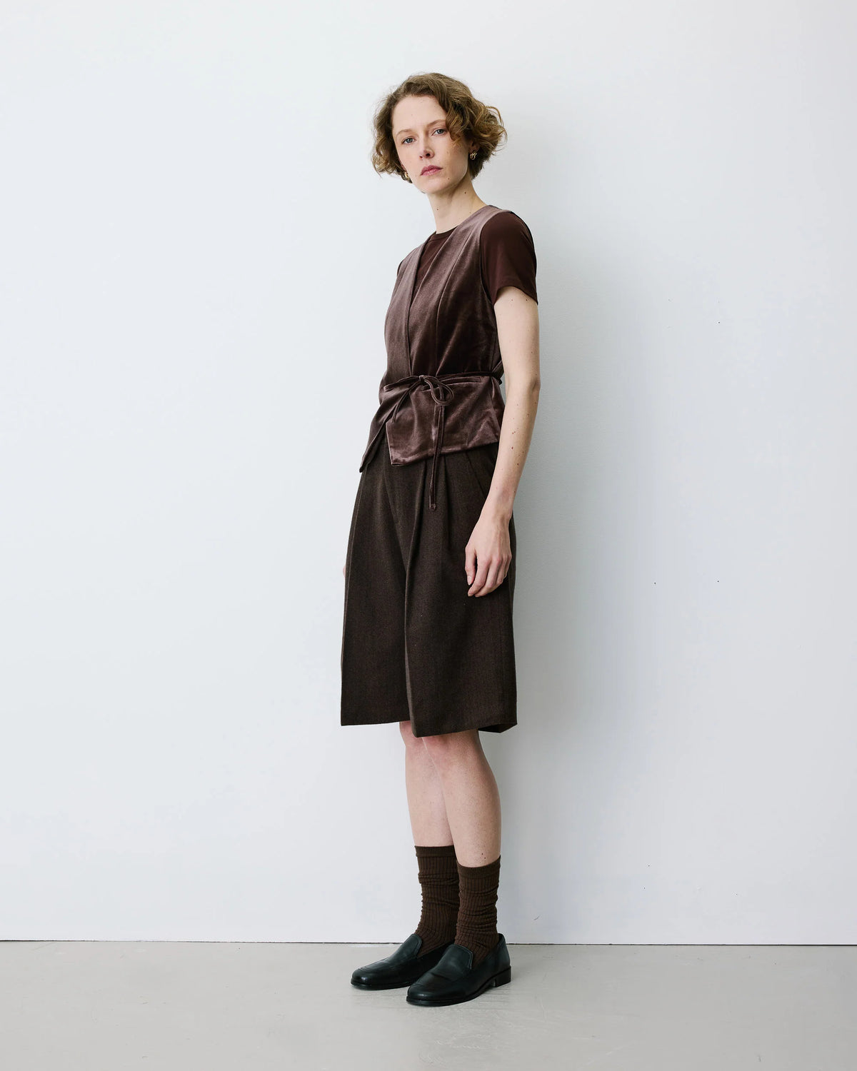 A model stands in profile wearing a dark brown short-sleeve dress with a satin-like textured bodice, a tied waist detail, and a straight skirt, paired with dark brown socks and black shoes, against a plain white studio wall.